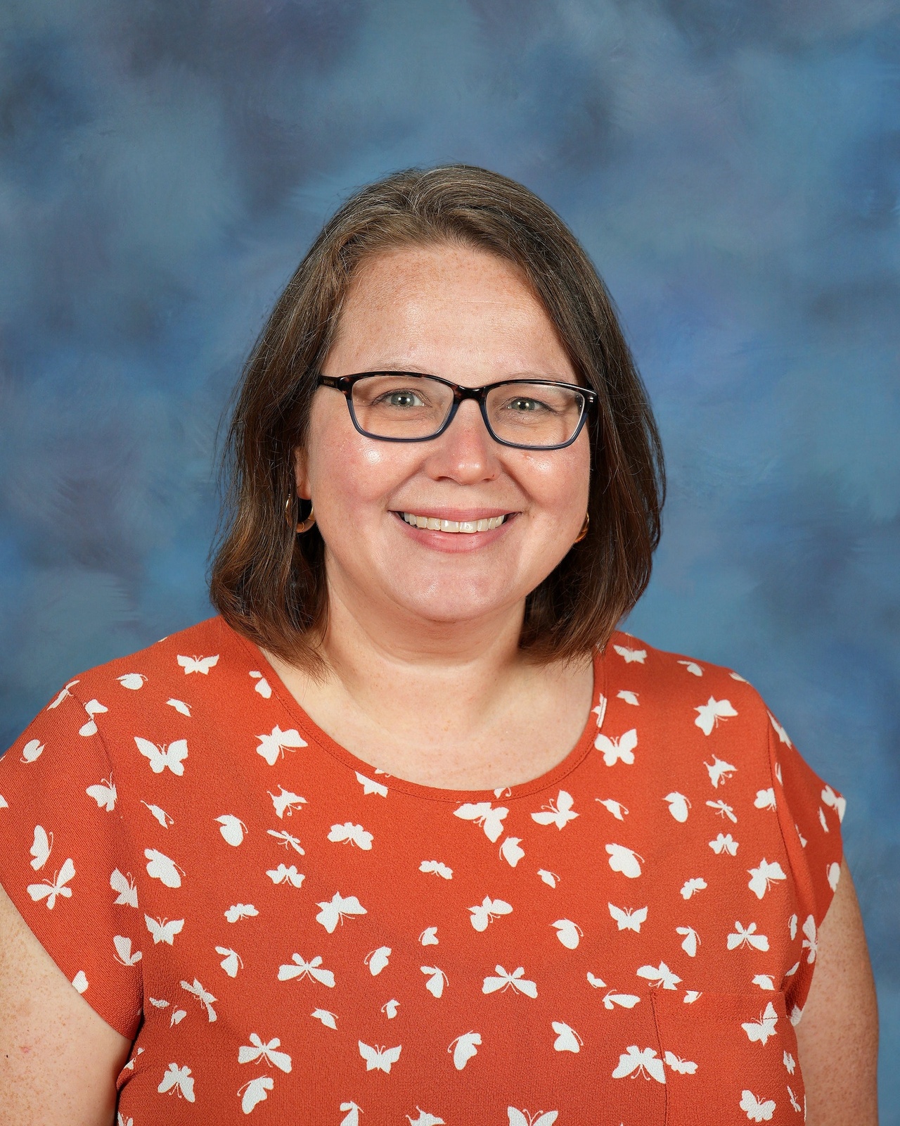 A smiling person with glasses and shoulder-length hair, wearing an orange top with white butterflies, against a blue, softly textured background.